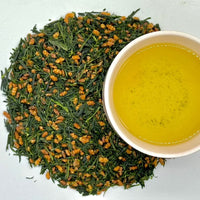 Loose leaf green tea mixed with roasted brown rice, with a cup of brewed tea beside it.