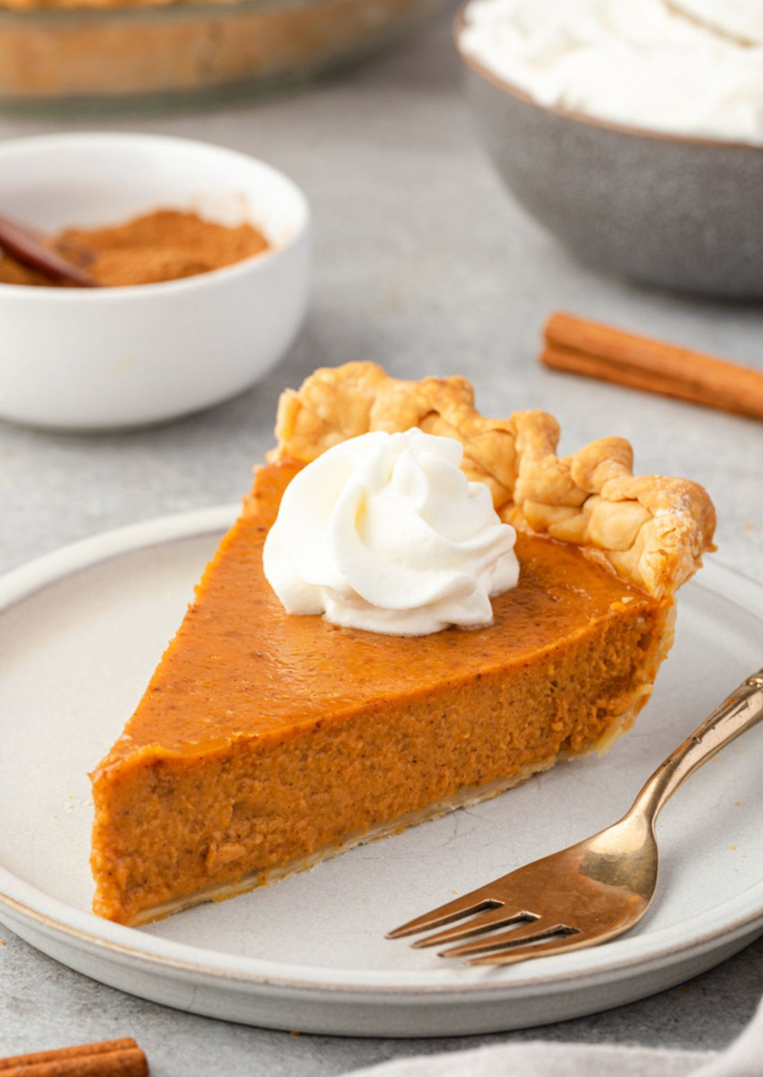 Slice of pumpkin pie with whipped cream on a plate, surrounded by cinnamon sticks and small bowls.