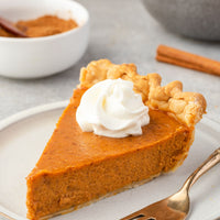 Slice of pumpkin pie with whipped cream on a plate, surrounded by cinnamon sticks and small bowls.