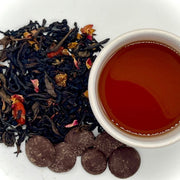 A box of tea and chocolates with Winter Palace Tea leaves visible along with a cup of brewed tea and vegan dark chocolate drops.