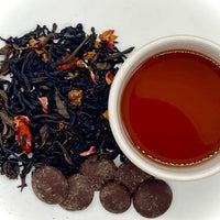 A box of tea and chocolates with Winter Palace Tea leaves visible along with a cup of brewed tea and vegan dark chocolate drops.
